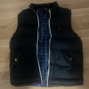 Svart dunväst från Polo Ralph Lauren - Säljer en snygg svart dunväst från Polo Ralph Lauren i storlek M. Västen har en dragkedja framtill och två fickor med knappar. Insidan är fodrad med ett blått och vitt rutigt mönster. Perfekt för höst och vinter! Dm mig vid intresse💋