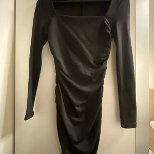 Svart långärmad klänning - Snygg svart klänning med långa ärmar och en elegant drapering framtill. Perfekt för festliga tillfällen. Materialet ser ut att vara mjukt och stretchigt, vilket ger en bekväm passform. Den har en stilren och tidlös design som passar bra till både höst och vinter.
