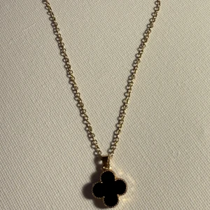Guldhalsband med fyrklöverhänge - Säljer ett elegant guldhalsband med ett svart fyrklöverformat hänge. Kedjan är tunn och stilren, perfekt för både vardag och fest. Hänget har en glansig finish som ger en lyxig känsla. Ett måste för smyckesälskare som vill ha något unikt och tidlöst.