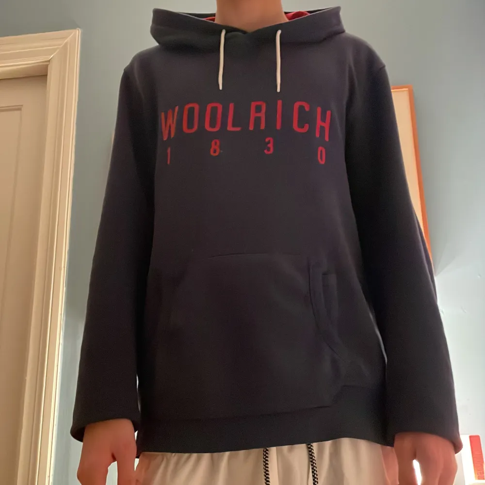 Säljer en snygg mörkblå hoodie från Woolrich med röd text på framsidan. Den har en  ficka fram och en  huva med vita snören. Perfekt för kyliga dagar och passar till det mesta. Använd ett fåtal gånger, så den är i bra skick. . Tröjor & Koftor.