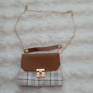 Väska - Säljer en superfin axelväska med rutigt mönster och brunt lock. Väskan har en elegant guldkedja och ett snyggt spänne framtill. Perfekt för både vardag och fest! 👜✨