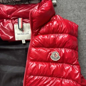 Röd dunväst från Moncler - Säljer en snygg röd dunväst från Moncler i modellen gui, äkta såklart. Västen är i storlek 3 vilket kan översättas som m-l. Västen är i mycket bra skick, förutom ena fickan som är lite sydd vid ett ställe. Är öppen för prisförslag! Det är bara att skriva för fler bilder eller frågor!