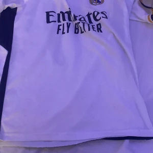 Vit Real Madrid fotbollströja - Säljer en vit Real Madrid fotbollströja med korta ärmar och snygga detaljer i guld och svart. Tröjan har 'Emirates Fly Better' tryckt på framsidan och 'Ronaldo 7' på baksidan. Perfekt för alla fans! 🏆