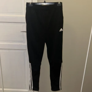 Svarta träningsbyxor från Adidas - Säljer ett par svarta träningsbyxor från Adidas i storlek 13-14 år. Byxorna har de klassiska tre vita ränderna längs sidorna och en liten Adidas-logga på låret. De är tillverkade i Primegreen-material, vilket är hållbart och perfekt för träning. Passar både för gymmet och löprundan!