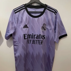 Real Madrid fotbollströja - Säljer en snygg lila Real Madrid tröja från Adidas i storlek S, passar även M. Skick 9/10. Tröjan är äkta. Den har korta ärmar och ett coolt mönster med svarta detaljer. På framsidan finns klubbens emblem och sponsorlogga, och på baksidan står det 'Benzema 9'. Perfekt för alla fans! 💜Bara att höra av sig vid minsta fundering!