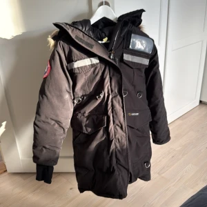 Svart Canada Goose  vinterjacka med pälsdetaljer - Säljer en svart vinterjacka från Canada Goose i mycket bra skick. Jackan har en varm pälsfodrad huva och flera praktiska fickor. Perfekt för kalla vinterdagar! Den har långa ärmar och en snygg passform. Märkeslogga på ärmen ger en extra touch. Jackan är väl använd, det finns defekter de sista bilden. 