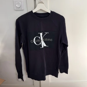 Svart tröja från Calvin Klein Jeans - Säljer en snygg svart tröja från Calvin Klein Jeans i storlek M. Tröjan har en stor logga på framsidan och är perfekt för både vardag och fest. Den är i mycket bra skick och har långa ärmar med ribbade muddar.