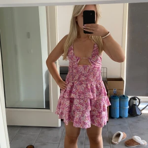 Rosa blommig klänning - Säljer en superfin rosa blommig klänning med volanger och öppen rygg. Den är ärmlös och perfekt för sommaren. Klänningen har en söt knytning framtill och är i kort modell. Perfekt för en somrig dag eller fest! 🌸