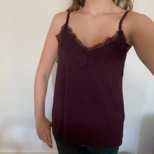 Vinröd linne med spetsdetaljer - Säljer ett snyggt vinrött linne med spetsdetaljer vid halsringningen. Perfekt för både vardag och fest! Linnet har smala axelband och en lös passform som gör det bekvämt att bära. Passar bra till jeans eller kjol för en stilren look.