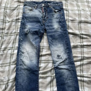 Blå jeans från Dsquared2 - Säljer ett par snygga blå jeans från Dsquared2 i storlek 44. De har en cool slitning och en patch med ett rött lönnlöv på bakfickan. Perfekta för en avslappnad stil med en touch av edge. Passar bra till både vardag och fest!