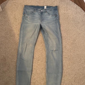 Ljusblå jeansbyxor - Säljer ett par ljusblå jeansbyxor i bra skick. De har en snygg slitning på ena benet och klassisk design. Perfekta för en avslappnad vardagslook. Passar bra till både sneakers och boots!
