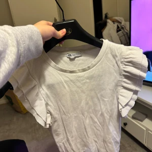 Vit topp med volangärmar från Zara - Säljer en söt vit topp från Zara med volangdetaljer på ärmarna. Perfekt för både vardag och fest! Toppen är i ett mjukt och bekvämt material, vilket gör den skön att bära hela dagen. I storlek M men passar även S