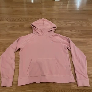 Rosa hoodie från GANT - Säljer en superfin rosa hoodie från GANT. Den är i bra skick och har en klassisk känguruficka framtill. Perfekt för mysiga dagar eller en avslappnad stil. Hoodien har långa ärmar och en bekväm passform. Perfekt för höst och vår! Orgilalpris 800kr