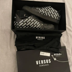 Svarta sneakers från Versus Versace - Säljer ett par limiterade svart/vita sneakers från Versus Versace i storlek 41 (passar även 42). Skorna har ett coolt mönster med 'VERSUS' tryckt över hela ytan. De är i bra skick och kommer med originalförpackning, tags och dustbag. Perfekta för att ge din outfit en lyxig touch!