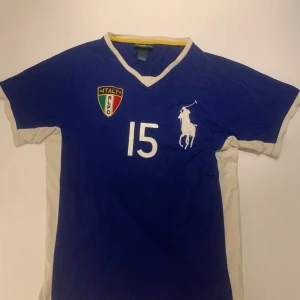 Blå fotbollströja från Ralph Lauren - Säljer en snygg blå fotbollströja från Ralph Lauren med nummer 15 på framsidan. Tröjan har korta ärmar och ett märke med texten 'ITALY R.L.F.C'. Den är i bra skick och har vita detaljer vid ärmslut och krage. Perfekt för sportiga tillfällen eller som en cool vardagströja!