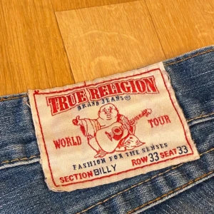 Blå jeans från True Religion - Säljer ett par blå True Religion jeans i bra skick. De har en cool slitning och en klassisk femficksdesign. Jeansen är i en straight stil. Perfekta för en avslappnad look!
