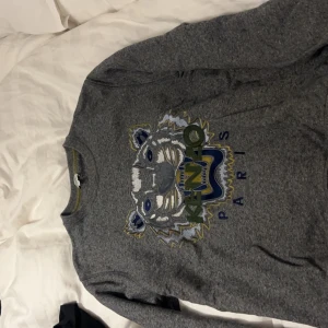 Grå sweatshirt från Kenzo - Säljer en grå sweatshirt från Kenzo med ett coolt tigertryck på framsidan. Tröjan har långa ärmar och är perfekt för både höst och vinter. Den är i bra skick och superbekväm att ha på sig. Passar perfekt till vardags eller en avslappnad dag ute.