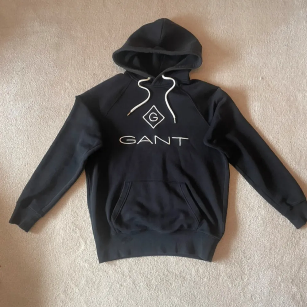 Säljer en snygg hoodie från GANT i storlek S. Den har en klassisk design med GANT-loggan broderad på framsidan och en praktisk magficka. Perfekt för både höst och vinter, och den är i mycket bra skick. Huvan har vita dragsnören för justering. Passar perfekt för en casual look!. Tröjor & Koftor.