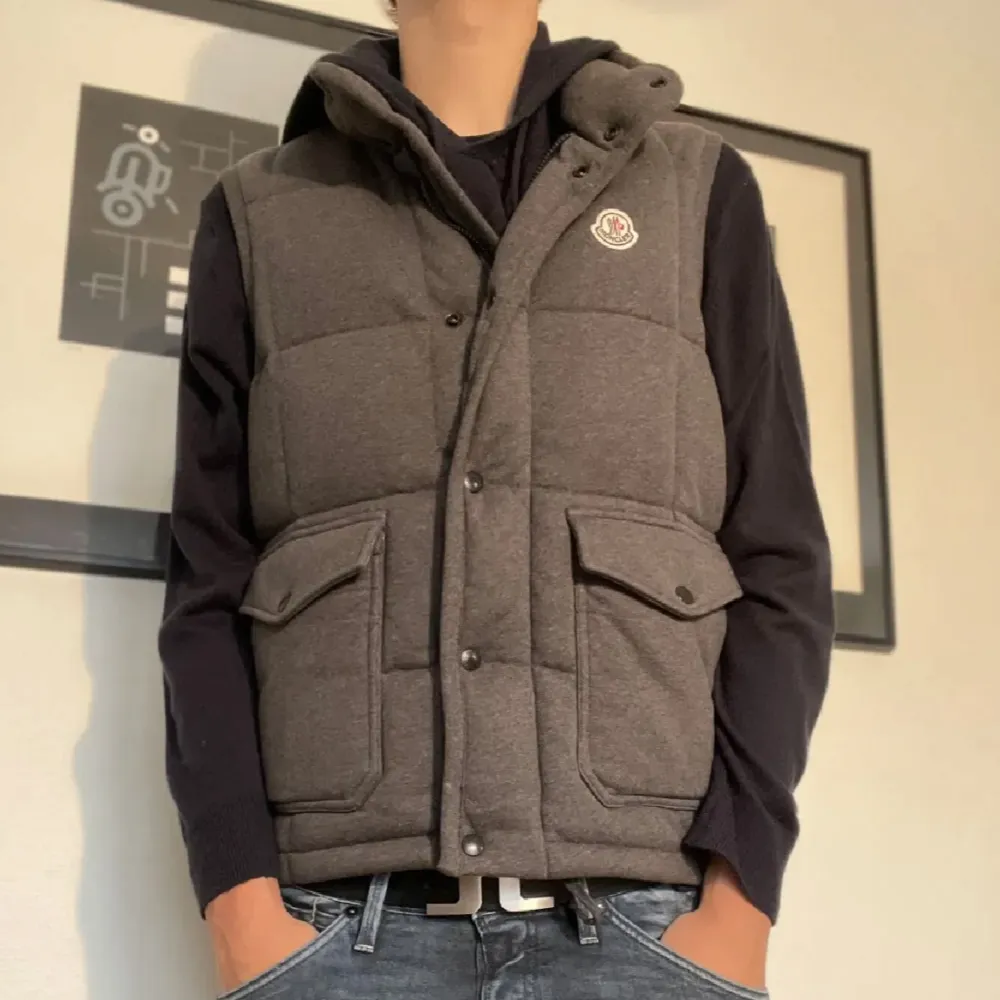 Säljer en snygg grå dunväst från Moncler i mycket bra skick. Västen har en quiltad design med knappar och två stora fickor framtill. Perfekt för höst och vinter, och den ger en stilren look. Passar bra till både vardag och fest. Storlek L men passar M också!. Jackor.