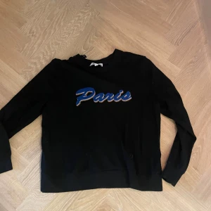 Svart tröja med Paris-tryck - Säljer en svart tröja med ett coolt blått och guldigt Paris-tryck på framsidan. Tröjan har långa ärmar och är perfekt för en avslappnad stil. Den är i bra skick och passar perfekt till höst- och vårgarderoben.