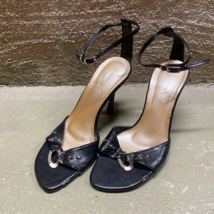 Svarta klackskor med detalj - Snygga svarta klackskor i skinn med en elegant rosett framtill. De har en smal rem runt ankeln och en stilren klack. Perfekta för festliga tillfällen eller en utekväll. Skorna är i bra skick och redo att användas för att ge din outfit det lilla extra.