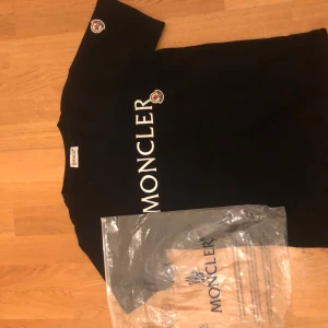 Svart t-shirt från Moncler - Säljer en svart t-shirt från Moncler i nytt skick. Den har en stor vit logga på framsidan och ett broderat märke på ärmen. Perfekt för en stilren och avslappnad look. Passar både till vardags och fest. Storlek XS men passar även S. pris kan diskuteras vid en snabb affär 💨