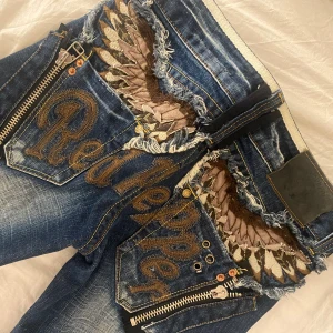 Blå jeans med broderade detaljer från Red Pepper - Säljer ett par unika blå jeans från Red Pepper med coola broderade detaljer och slitningar. De har dragkedjor och en snygg design på bakfickorna. Perfekta för att sticka ut i mängden! Säljer pga att de är lite för korta på mig som är 167
