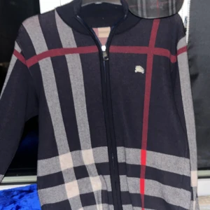 Rutig kofta från Burberry - Säljer en snygg rutig kofta från Burberry i mörkblått med röda och beige detaljer. Den har en dragkedja framtill och ett litet broderat emblem på bröstet. Perfekt för höst och vinter, både stilren och bekväm. Passar lika bra till vardags som till fest! Kom dm för mer information och bilder/frågor 