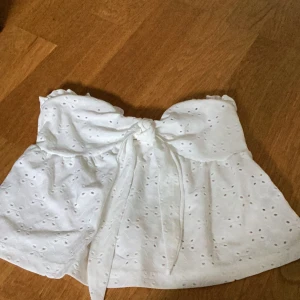 Vit axelbandslös topp med knyt - Säljer en superfin vit axelbandslös topp med knyt framtill. Den har ett sött broderat mönster och är perfekt för sommardagar. Toppen är i ett lätt och luftigt material, vilket gör den idealisk för varma väder. Från Shein