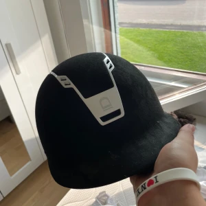 Svart ridhjälm från Equipage - Säljer en svart ridhjälm från Equipage. Hjälmen har en stilren design med en vit detalj framtill. Den är utrustad med justerbart spänne för perfekt passform och har en mjuk insida för extra komfort. Perfekt för ridning och säkerhet!