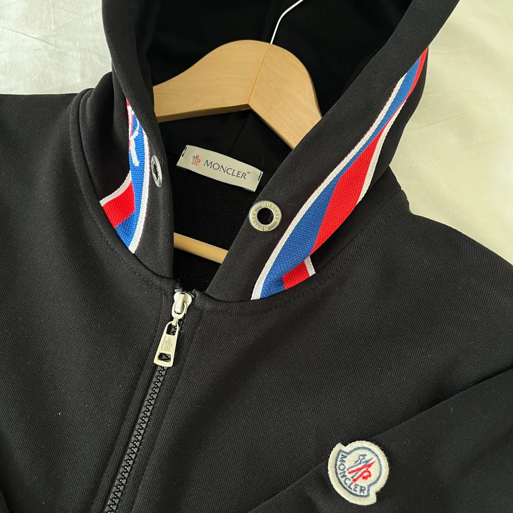 Säljer en snygg svart hoodie från Moncler. Den har en dragkedja framtill och en huva med röd och blå randig detalj. Moncler-loggan sitter på bröstet. Perfekt för en casual look eller när du vill hålla det stilrent och bekvämt.. Hoodies.