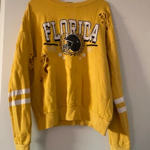 Gul tröja från Gina Tricot med Florida-tryck - Säljer en gul tröja från Gina Tricot med ett coolt Florida-tryck och en amerikansk fotbollshjälm på framsidan. Tröjan har långa ärmar med vita ränder och är lite sliten med hål för en edgy look. Perfekt för en avslappnad stil! Hör gärna av er vid frågor eller fler bilder💕