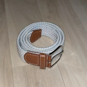 Vita flätat bälte med bruna detaljer - Säljer ett snyggt vit flätat bälte med bruna läderdetaljer. Perfekt för att ge en stilren touch till vilken outfit som helst. Frågor och funderingar? Hör bara av dig.