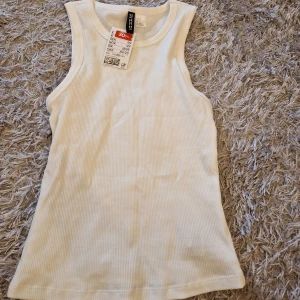 Vit ribbad linnetopp från H&M Divided - Säljer en snygg vit ribbad linnetopp från H&M. Perfekt för sommaren med sin ärmlösa design och bekväma passform. Toppen är i nyskick och har en klassisk stil som passar till det mesta. Perfekt för både vardag och fest!