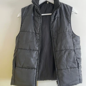 Grå quiltad dunväst - Säljer en snygg och praktisk svart dunväst med quiltat mönster. Västen har en dragkedja framtill och är perfekt för kyligare dagar. Den är i bra skick och passar både till vardags och för utomhusaktiviteter.