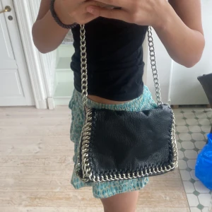 Svart axelväska från Tiamo - Säljer en snygg svart axelväska från Tiamo. Väskan är i skinn och har en cool kedjedetalj runt kanterna. Den har en dragkedja på baksidan för extra förvaring. 