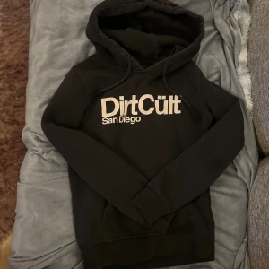 Svart hoodie från DirtCult - Säljer en svart hoodie från DirtCult med texten 'San Diego' tryckt på framsidan. Den är superbekväm och perfekt för kyliga dagar. Hoodien har en klassisk passform med långa ärmar och en praktisk ficka framtill. Perfekt för både vardag och chill! 🖤