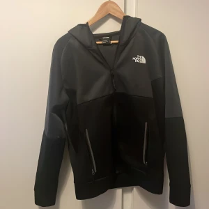 Svart och grå jacka från The North Face - Säljer en snygg svart och grå kofta från The North Face. Den har en dragkedja framtill och två praktiska fickor med dragkedjor. Perfekt för höst och vår, med en bekväm passform och huva för extra skydd mot väder och vind.