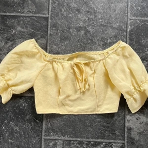 Gul offshoulder topp - Säljer en superfin gul offshoulder topp med korta puffärmar och knytning framtill. Perfekt för sommardagar och ger en söt och romantisk look. Toppen är croppad och gjord i ett lätt och bekvämt material.