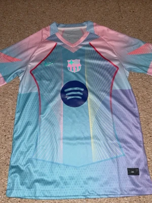 Barcelona pastellfärgad fotbollströja - Säljer en Barcelona fotbollströja i pastellfärger med blå, rosa och lila toner. Tröjan har korta ärmar, V-ringning och tryckta detaljer som klubbmärke och sponsorlogga på bröstet. Materialet är lätt och andas, perfekt för träning eller match.