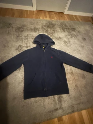 Mörkblå hoodie med dragkedja - Mörkblå hoodie med dragkedja och huva. Klassisk design med två fickor framtill och en liten röd logga på bröstet. Tillverkad i mjukt material som känns skönt mot huden. Perfekt för en avslappnad och stilren look.
