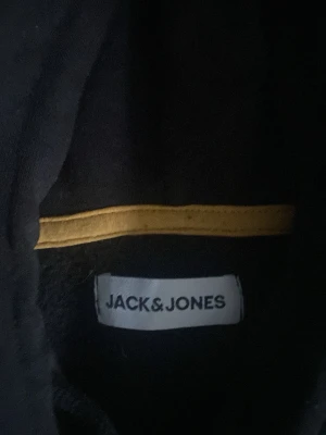 Svart hoodie från Jack & Jones - Snygg svart hoodie från Jack & Jones med vit grafisk text på framsidan. Klassisk huva med dragsko och ribbade muddar vid ärmar och nederkant. Perfekt för en avslappnad och trendig stil.