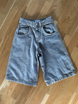 Grå-brun loose denimshorts - Säljer ett par grå-bruna denimshorts med loose fit och klassisk femficksdesign. Shortsen har knappgylf och bälteshällor, samt raka ben som slutar strax under knät. Perfekta för en avslappnad stil. 