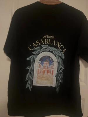 Svart t-shirt från Casablanca - Svart t-shirt från Casablanca med tryck på ryggen som föreställer ett fönster med utsikt mot en byggnad och natthimmel. Texten 'Avenida Casablanca' ovanför motivet. Klassisk passform och rund hals.