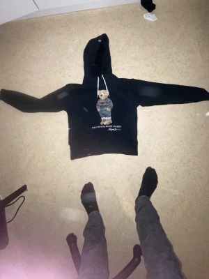 Ralph lauren hoodie - Svart hoodie från Polo Ralph Lauren med en ikonisk björn på bröstet. Hoodien har huva med vita snören och en avslappnad passform. Perfekt för dig som gillar streetwear och vill sticka ut med en klassisk design.