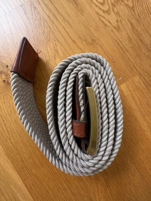 Beige flätat bälte med läderdetaljer - Snyggt beige flätat bälte med bruna läderdetaljer och ett metallspänne i guldton. Bältet har en flexibel och stretchig design som gör det bekvämt att bära. Passar perfekt till jeans eller chinos för en avslappnad men stilren look.