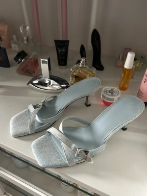 Ljusblå sandaler med smal klack - Snygga ljusblå sandaler med smal klack och fyrkantig tå. Skorna har två tunna remmar över foten, varav en med silverfärgat spänne. Materialet ser ut att vara textil med en mjuk finish och innersulan är matchande ljusblå. Perfekta för dig som gillar stilrena detaljer.