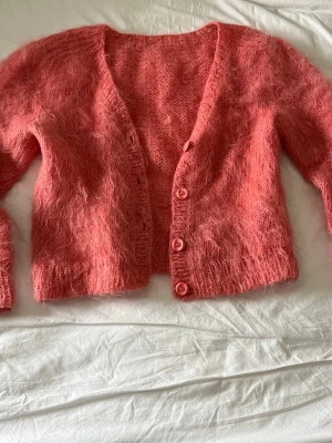 Rosa fluffig kofta med knappar - Supermjuk och fluffig rosa kofta med v-ringning och tre matchande knappar framtill. Koftan är stickad i ett lurvigt material och har långa ärmar. Perfekt för dig som gillar mysiga och trendiga plagg.
