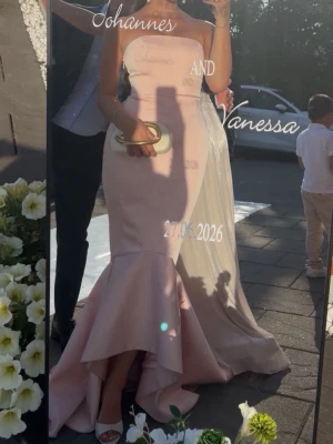 Ljusrosa axelbandslös långklänning - Elegant ljusrosa långklänning med axelbandslös design och figurnära passform. Klänningen har en volangdetalj nedtill som ger en dramatisk effekt. Perfekt för bal eller festliga tillfällen. Tillverkad i ett slätt material som ger en lyxig känsla.