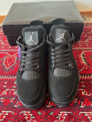 Svarta Air Jordan 4 Retro sneakers - Snygga helsvarta Air Jordan 4 Retro sneakers med klassisk siluett och meshdetaljer på sidorna. Skorna har svarta skosnören, Jumpman-logga på plösen och robust sula. Materialet är en mix av mocka och syntet. Perfekta för dig som gillar streetwear och sportig stil.